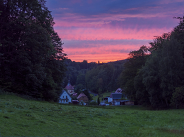 Roter Abendhimmel über kleinem Dorf in Bergschlucht mit Kirche, Wiese und Wäldern, Lückendorf, Zittauer Gebirge, Sachsen, Deutschland