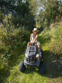 Frau fährt Rasenmäher, Rasentraktor, mit Strohhut und Sonnenbrille, im Garten bei der Arbeit, Deutschland