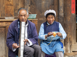Altes chinesisches Ehepaar sitzt vor Holzhaus, Frau häkelt, Mann raucht Bong, Shaxi, Yunnan, China