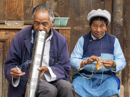 Altes chinesisches Ehepaar sitzt vor Holzhaus, Frau häkelt, Mann raucht Bong, Shaxi, Yunnan, China